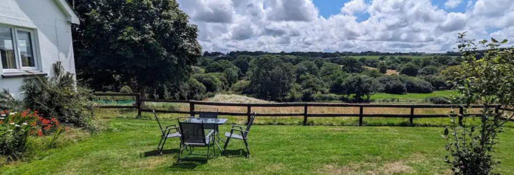 View in garden with seating a table, clouds, trees, to the left a white house wall and windows