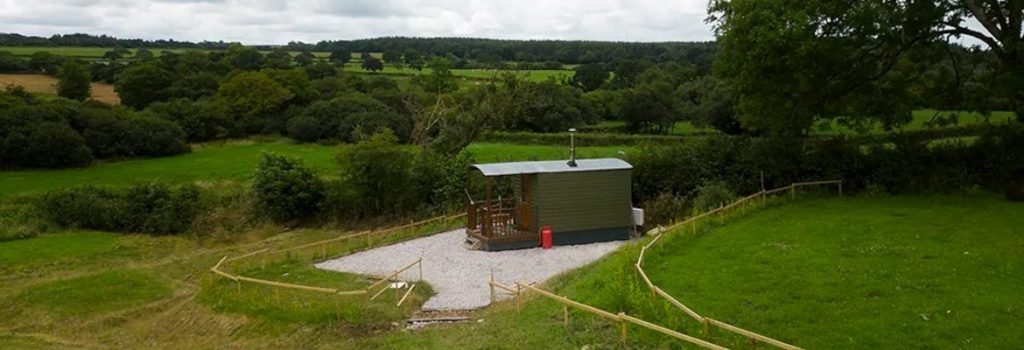 Shepherds hut on a hill, on a gravel base. Surrounded by wooden fencing and grass - GALLERY